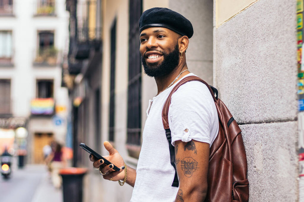 Stilvoller schwarzer Mann mit Baskenmütze und Lederrucksack, der im Sommer mit dem Smartphone auf einer Straße in der Stadt spaziert - Foto, Bild