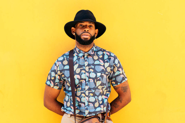 Portret van een stijlvolle zwarte man met hoed en alternatieve outfit over gele achtergrond - Foto, afbeelding
