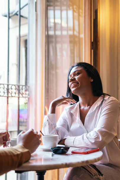 Close-up van een mooie Latijnse vrouw in casual kleding glimlachend terwijl ze praat met iemand in een koffieshop. Ze kijkt opgewonden en attent naar haar partner. Haar ogen geven de zenuwen en opwinding van een eerste afspraakje weer. - Foto, afbeelding