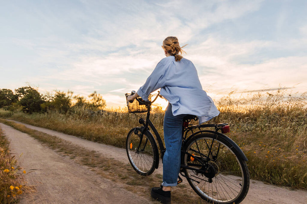 вид сзади блондинки на велосипеде в поле на закате - Фото, изображение