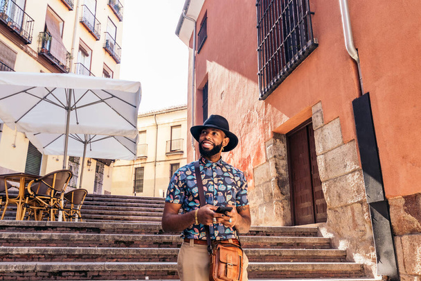 Стильний чорний чоловік з капелюхом, що ходить по міській вулиці влітку, використовуючи смартфон - Фото, зображення