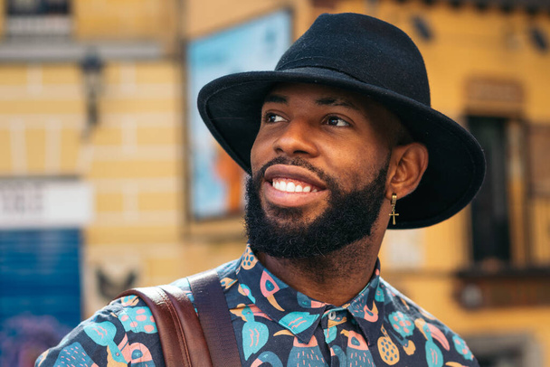 Elegante uomo di colore con look hipster alternativi passeggiando per le strade di una città in una giornata estiva soleggiata. sorride felicemente - Foto, immagini