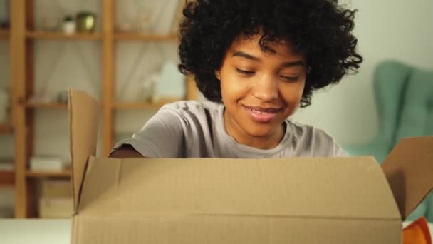 Μια Αφρικάνα ξεπακετάρει ψάχνοντας στο κουτί. Ευτυχισμένη γυναίκα ανοίγει χαρτόκουτο. Γυναίκα να πάρει δέμα κοιτάζοντας παραδοθέντα προϊόντα. Ικανοποιημένος πελάτης θετική ανατροφοδότηση γρήγορη παραδώσει - Πλάνα, βίντεο
