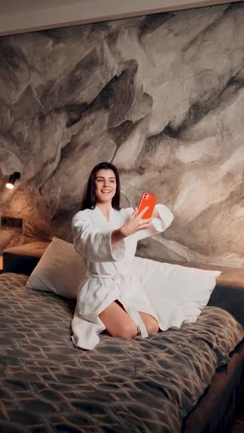 Młoda kobieta w szlafroku robi sobie rano selfie w łóżku w hotelu. Koncepcja wakacji hotelowych. Pionowe wideo - Materiał filmowy, wideo