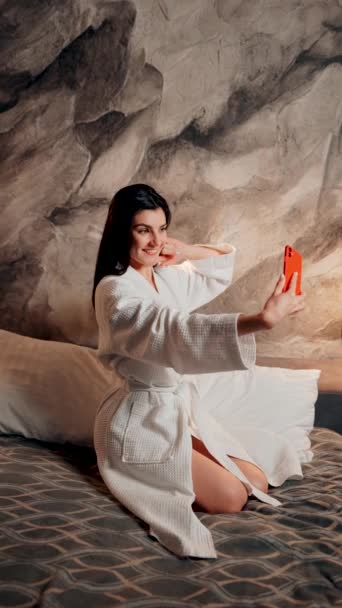 Una mujer joven con albornoz se toma una selfie en la cama por la mañana en un hotel. Concepto de vacaciones del hotel. Vídeo vertical - Metraje, vídeo