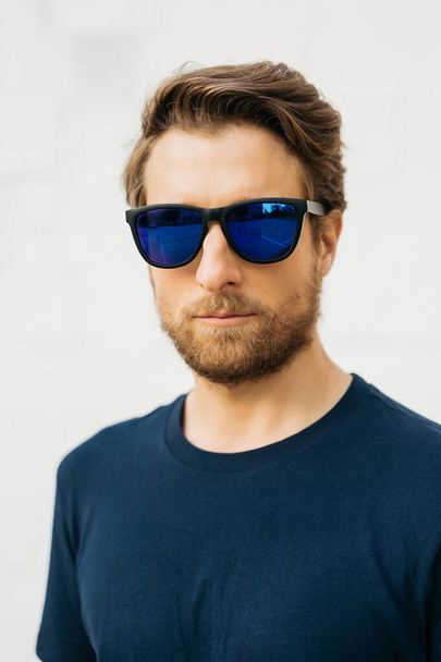 Ritratto di un uomo che si erge fiducioso contro una parete bianca, indossa occhiali da sole riflettenti e una t-shirt blu, trasudando un comportamento fresco e composto. - Foto, immagini