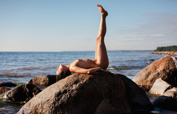 Eine junge schöne Frau posiert nackt zwischen großen Felsen am Ufer des Meeres. Jugend- und Nacktkonzept. - Foto, Bild