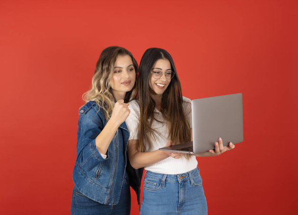 Две подружки IT-женщины держат ноутбук на фоне красной студии. Совместная работа на компьютере. Носите джинсовую одежду, выглядящую как экран, сделайте жест победителя, сжав кулак. Копирование пространства. - Фото, изображение