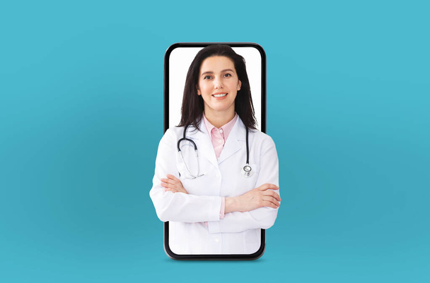 Γυναίκα γιατρός φορώντας ένα λευκό παλτό και στηθοσκόπιο διεξάγει μια εικονική διαβούλευση. Η τεχνολογία τηλεϊατρικής, με την εμφάνισή της μέσα από μια οθόνη smartphone σε μπλε φόντο. - Φωτογραφία, εικόνα