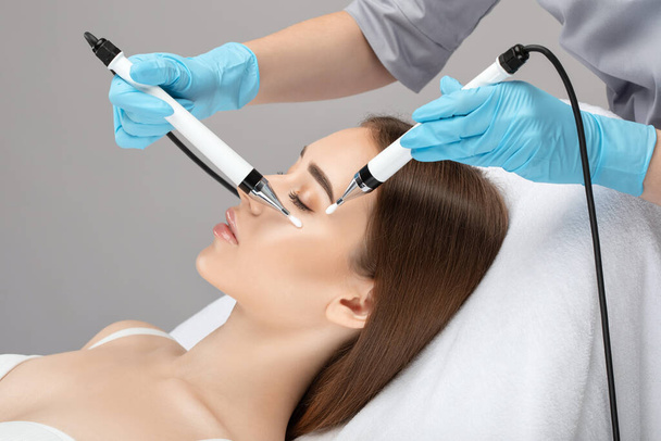 A doctor cosmetologist makes a microcurrent facial therapy to a young woman with a device in a beauty wellness salon.Cosmetology and professional skin care. - Photo, Image