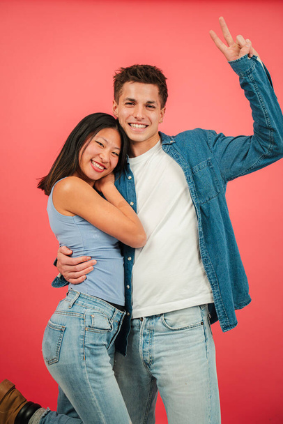 Κάθετη Νεαρό πολυφυλετικό ζευγάρι χαμογελά και αγκαλιάζει μπροστά από ένα φόντο στούντιο κόκκινο χρώμα. Δύο άνθρωποι σε μια ρομαντική και ευτυχισμένη στάση κοιτάζοντας την κάμερα μαζί. Υπέροχος φίλος και φίλη - Φωτογραφία, εικόνα