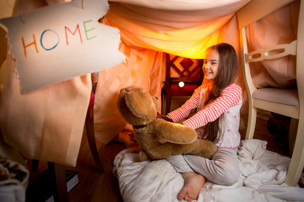 Χαριτωμένο χαμογελαστό κορίτσι παίζει με αρκουδάκι στο σπίτι κατασκευασμένο από κενό - Φωτογραφία, εικόνα