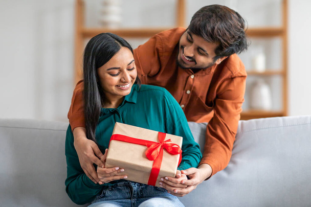 Indian man sitting on a couch is handing a gift to a woman next to him. The woman appears surprised and delighted as she receives the gift - Foto, imagen