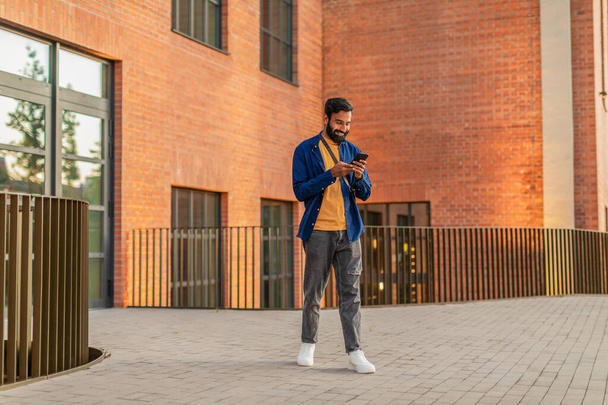 Indian man is standing in front of a brick building, focused on his cell phone screen. He appears engrossed in his device, with the urban backdrop of the building behind him. - Fotografie, Obrázek