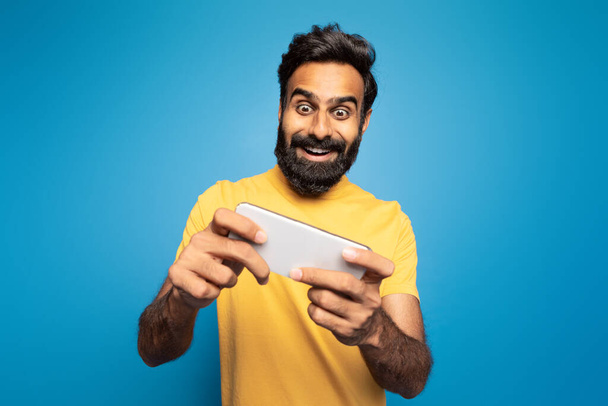 Um homem indiano alegre vestindo uma camisa amarela está entusiasticamente jogando um jogo para celular. Ele é definido contra um fundo azul vibrante, totalmente imerso no jogo. - Foto, Imagem