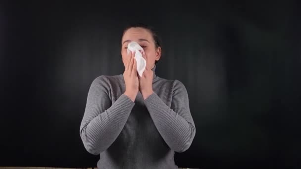 Muotokuva nuori nainen yllään harmaa villapaita puhaltaa nenän lautasliinaan mustalla taustalla sisätiloissa. Ruumiillinen reaktio aivastus aiheuttama sairaus, kylmä tai allergioita. Terveydenhuolto, hengitysvaikeudet - Materiaali, video