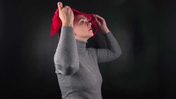 Een mooie jonge vrouw trekt een rode zakdoek aan, draagt een grijze trui met een ongedwongen uitstraling, haar doordringende blik straalt een scherpe maar zelfverzekerde houding uit tegen de minimale zwarte achtergrond. - Video
