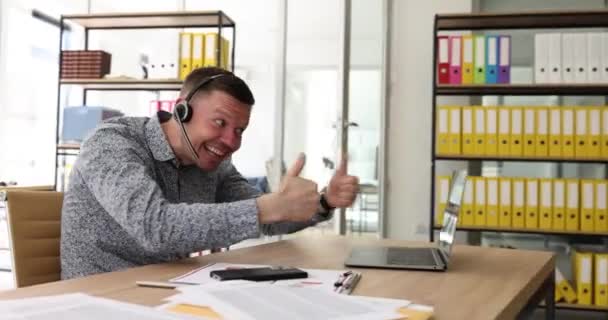 Een lachende zakenman in een koptelefoon kijkend naar een laptop scherm, kijkend naar een goede webinar die duimen opsteekt. Mood swings consulting client of het leren van talen op afstand tonen gebaar fak deelnemen aan - Video
