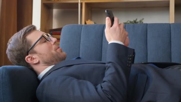 No vídeo, um homem de terno está em um sofá azul, olhando para o telefone, dormindo e tirando os óculos. Ele parece relaxado e focado em seu telefone enquanto descansa - Filmagem, Vídeo