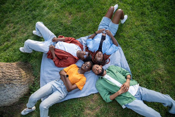 Gruppo di riflessivi rilassati giovani amici afroamericani sdraiati insieme su erba verde all'aperto. Vista dall'alto di quattro studenti neri che riposano sul plaid nel parco, godersi la natura e il silenzio. Amicizia. - Foto, immagini