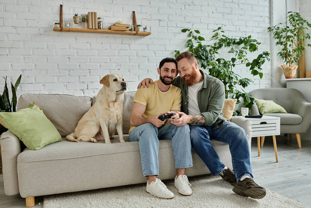 Δύο άντρες, ένα γκέι ζευγάρι, κάθονται σε έναν καναπέ παίζοντας βιντεοπαιχνίδι με το λαμπραντόρ σκύλο τους σε ένα άνετο σαλόνι. - Φωτογραφία, εικόνα