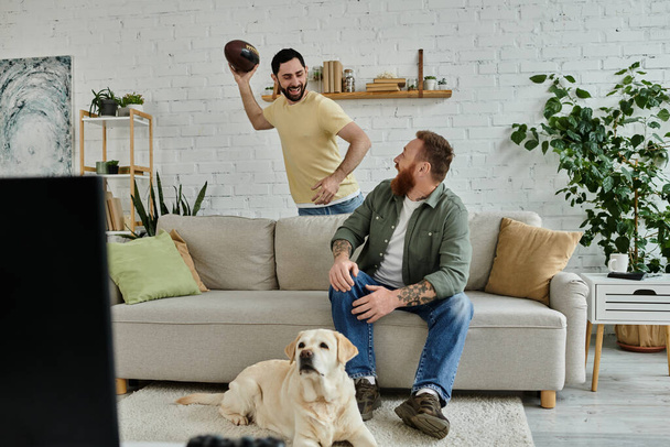 Ein Mann mit Bart sitzt mit einem Labrador auf einer Couch, beide in ein Sportspiel im Fernsehen vertieft. - Foto, Bild
