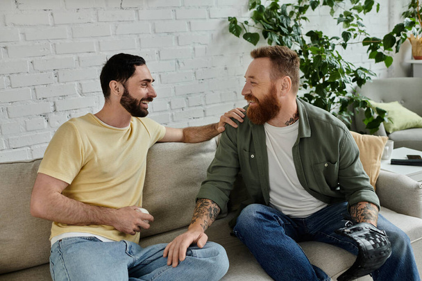 Two bearded men chat on a cozy couch watching a sports match, enjoying quality time together. - Photo, Image