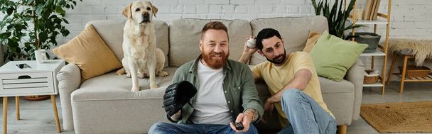 Dos hombres con barba descansando en un sofá, absortos en ver un partido deportivo con su perro Labrador a su lado. - Foto, Imagen