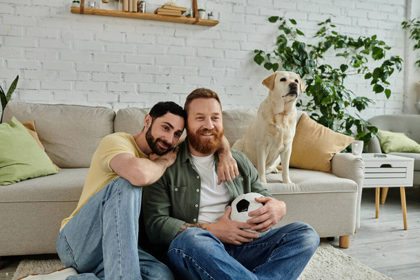 Двое бородатых мужчин сидят на диване, смотрят спортивный матч с собакой-лабрадором на заднем плане. - Фото, изображение