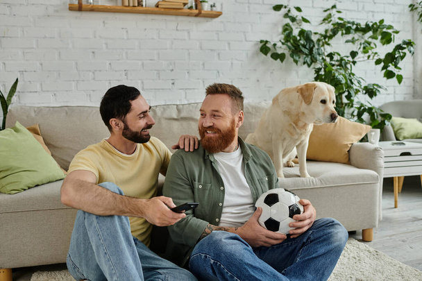 Dos hombres con una pelota de fútbol, descansando en un sofá con su perro labrador, absortos en un partido de deportes en la televisión. - Foto, Imagen
