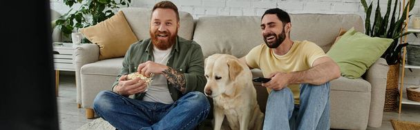 Zwei bärtige Männer sitzen mit einem Labrador auf einer Couch und genießen gemeinsam in einem gemütlichen Wohnzimmer den Komödienfilm. - Foto, Bild