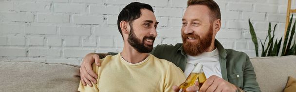 barbudo gay casal clinking cerveja garrafa e sentado no sofá no sala de estar. - Foto, Imagem