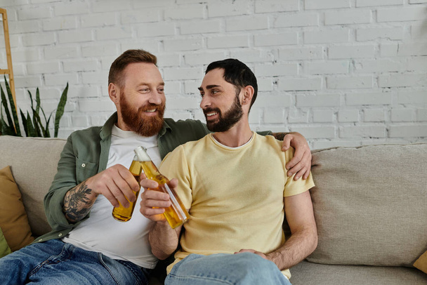 Bearded happy gay couple relaxes on sofa clinking beer bottles at home - Photo, Image