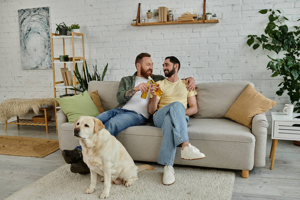 Two men, a Bearded gay couple, sit on a couch watching a sport match with their labrador dog by their side in the living room. - Photo, Image