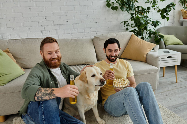 リビングルームのソファーでラブラドール犬と一緒にスポーツの試合を喜んで見ている2人のひげ付き男性. - 写真・画像
