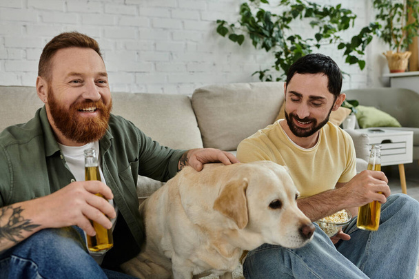 Deux hommes, un couple gay, se détendent sur un canapé en regardant un match de sport avec leur chien Labrador dans leur salon. - Photo, image