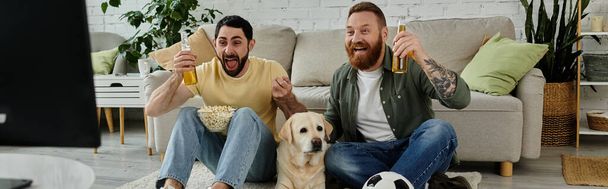 Una pareja gay y su Labrador sentados encima de un sofá, viendo un partido deportivo en la sala de estar. - Foto, Imagen