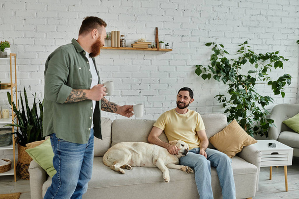 Deux hommes, un debout avec des tasses, un assis sur un canapé, dans un salon avec un chien labrador à proximité. - Photo, image