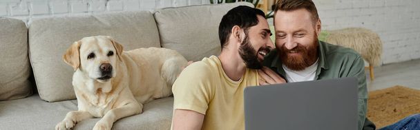 Γενειοφόρο ομοφυλόφιλο ζευγάρι και το Λαμπραντόρ σκυλί τους κάθονται σε έναν καναπέ, και οι δύο ασχολούνται με την προβολή περιεχομένου σε ένα φορητό υπολογιστή. - Φωτογραφία, εικόνα