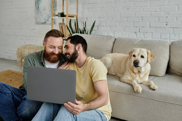 Bärtiger Mann und Labrador-Hund sitzen auf einer Couch, konzentriert auf einem Laptop-Bildschirm in einem gemütlichen Wohnzimmer. - Foto, Bild