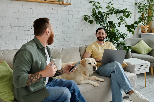Bärtiges schwules Paar sitzt auf einer Couch mit Labrador-Hund und arbeitet in einem gemütlichen Wohnzimmer an einem Laptop. - Foto, Bild
