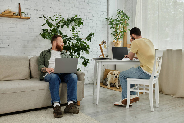 Un par de hombres están sentados encima de un sofá en una sala de estar, trabajando remotamente con un perro labrador cerca. - Foto, imagen