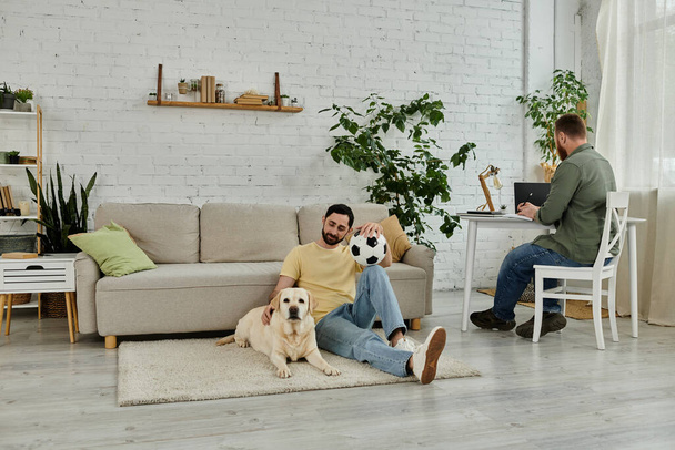 Bärtiger schwuler Mann sitzt auf dem Boden, zwei Labrador-Hunde an seiner Seite im gemütlichen Wohnzimmer, während er fernab arbeitet. - Foto, Bild