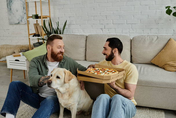 Zwei bärtige Männer sitzen auf dem Fußboden und essen Pizza, während ihr treuer Labrador-Hund in gemütlicher Wohnzimmeratmosphäre zu ihnen stößt.. - Foto, Bild