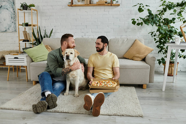 Deux hommes à la barbe se détendent sur un canapé avec une boîte de pizza, partageant une tranche tandis qu'un Labrador amical regarde. - Photo, image