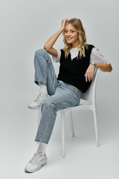 Una giovane donna elegante con i capelli biondi siede elegantemente su una sedia bianca su uno sfondo grigio. - Foto, immagini