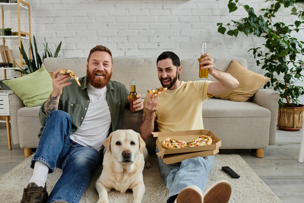 Two men with beards enjoy pizza and beer on a couch with their Labrador dog in a cozy living room setting. - Photo, image