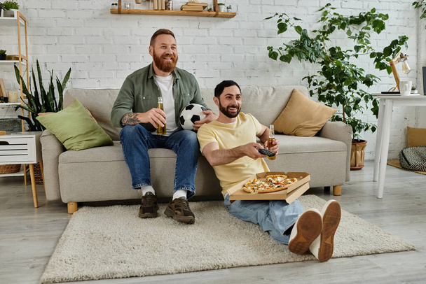 Dwóch brodatych mężczyzn relaksuje się na kanapie, dzieląc się pizzą i piwem w przytulnym salonie. - Zdjęcie, obraz