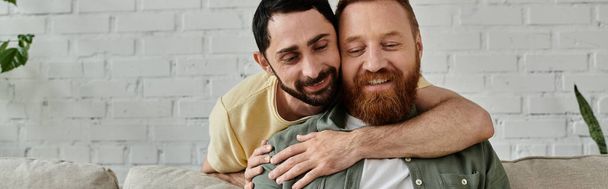 Двоє бородатих чоловіків обіймаються ласкаво на затишному дивані у своїй вітальні. - Фото, зображення