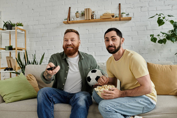 Twee bebaarde mannen zitten op een bank en houden een voetbal vast, genietend van quality time samen in hun gezellige woonkamer.. - Foto, afbeelding